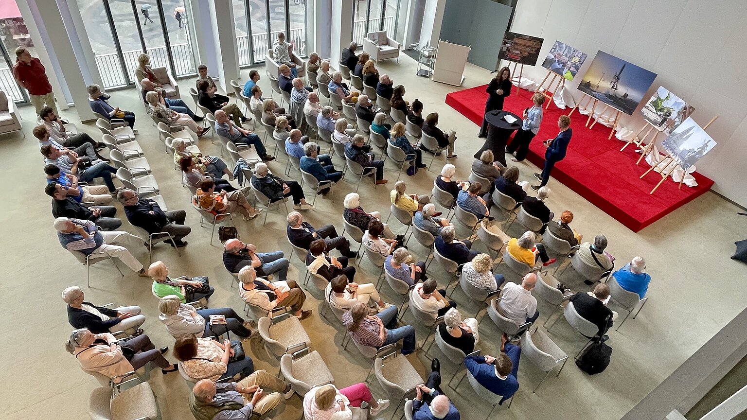 Festsaal im Haus der Bürgerschaft: Viele Menschen sitzen auf Stühlen vor einer Bühne. Auf der Bühne stehen Bilder. Drei Personen stehen vorne an einem Stehtisch. – 