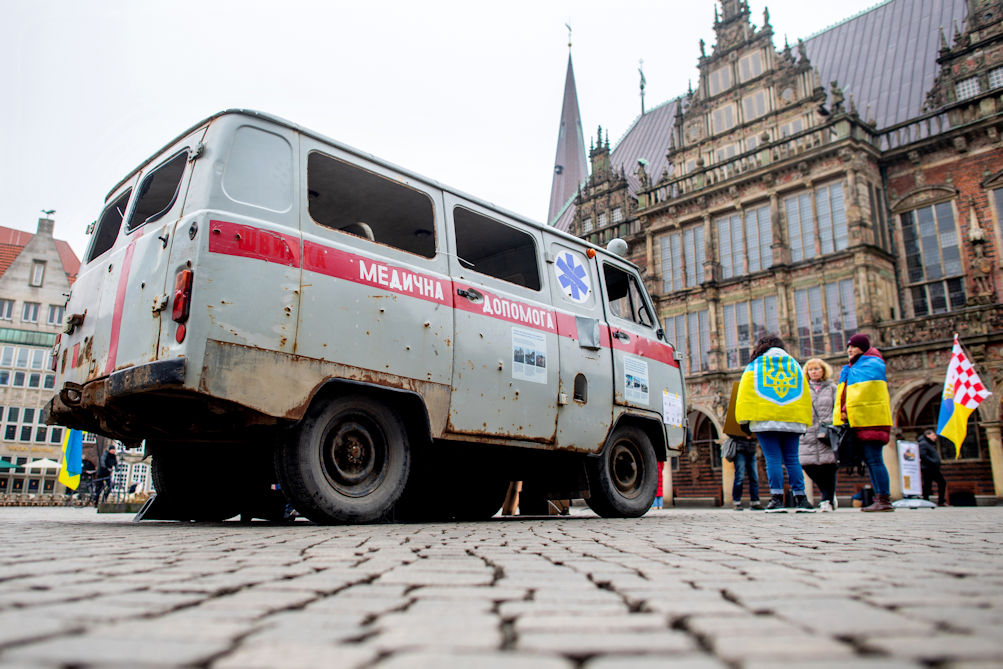 Ein ukrainischer Krankenwagen mit Einschusslöchern steht vor dem historischen Bremer Rathaus. Das Bild wurde in Froschperspektive aufgenommen. Im Hintergrund sind einige Demonstant*innen mit umgelegten Ukraine-Flaggen zu sehen. – Pressefoto des Jahres